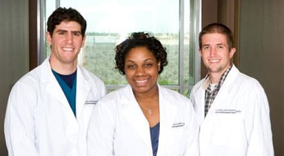 Three medical students wearing white lab coats smiling, posed for a picture.