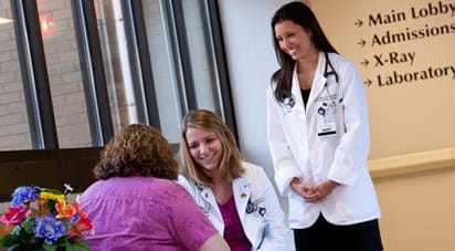 Female medical students wearing white lab coats, smiling while consulting with a patient.