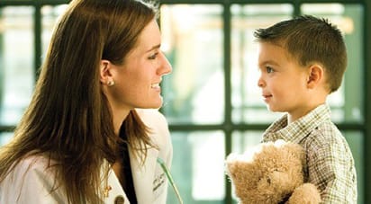 A young woman wearing a white lab coat, talking with a small boy holding a teddy bear.